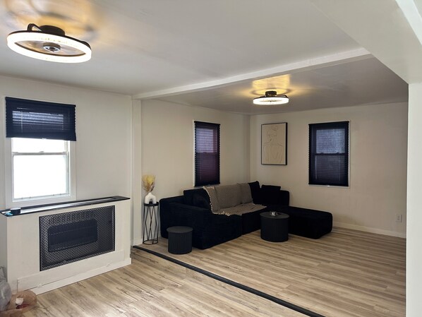A modern living room with a black sectional sofa, a small side table, and a radiator. There are three windows with blinds, a ceiling light fixture, and a framed artwork on the wall.