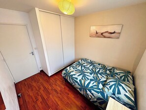 A bedroom with a wooden floor, a bed with a leaf-patterned bedspread, a white wardrobe, and a wall-mounted picture.