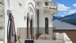 Eine Terrasse mit Gartenmöbeln, ein weißes Gebäude mit Verzierungsbögen und Blick auf Berge.