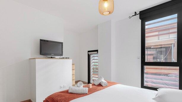 A modern hotel room with a bed, a television on a wooden stand, a window with a view of a building, and a red textured blanket. - Modern townhouse with parking (Toulouse)