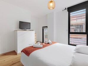 Et moderne hotelværelse med en seng, et fjernsyn på et træstel og et vindue med udsigt til en bygning, samt et rødt, struktureret tæppe.