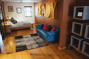 A cozy living room with a blue sofa, a bed, wooden flooring, and a 'READY' sign on the wall. - NO.1 Bakewell - New, modern one bedroom apartment with private parking. (Bakewell)