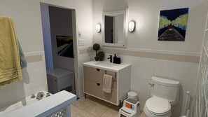Une salle de bain avec un lavabo blanc, un miroir et des toilettes, une petite plante en pot et un tableau accroché au mur.