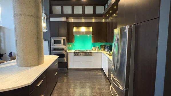 Une cuisine moderne avec des meubles de couleur sombre, un îlot central, un réfrigérateur en acier inoxydable et un pare-feu vert.