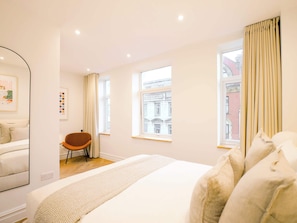 Ein Schlafzimmer mit einem Bett, einem Sessel, einem Spiegel und zwei Fenstern mit Vorhängen.