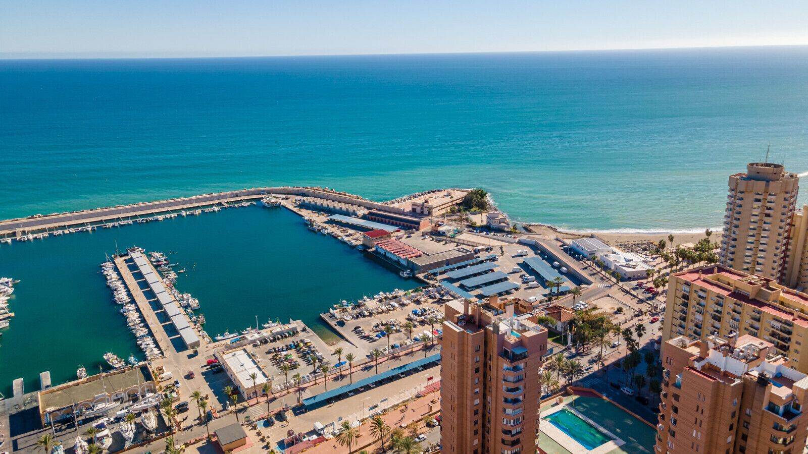 Vue aérienne d’une marina côtière, avec un quai, des bâtiments adjacents et une plage.