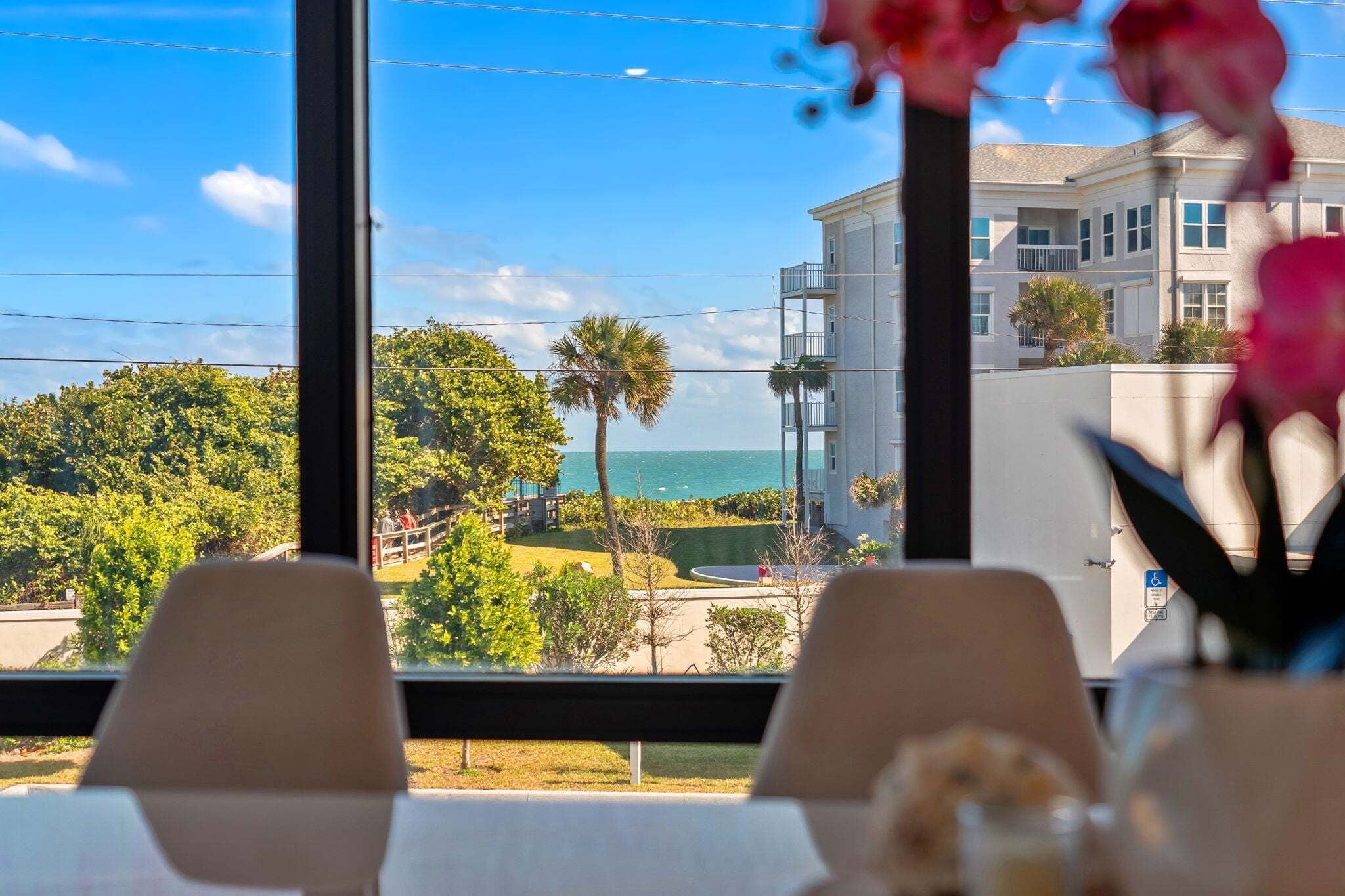 A view from a window with a table and chairs, overlooking a building with a balcony and a palm tree.