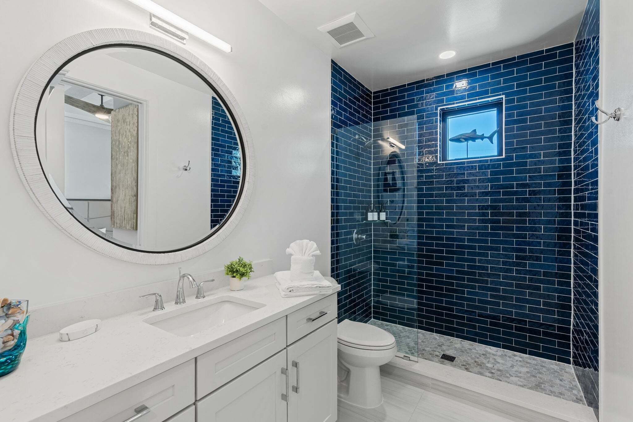 A bathroom with a white vanity, a round mirror, a blue-tiled shower wall, and a toilet.