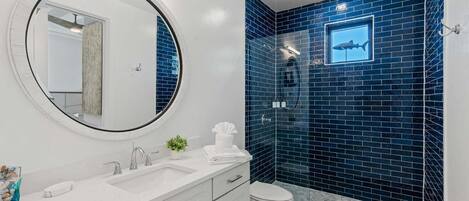 A bathroom with a white vanity, a round mirror, a blue-tiled shower wall, and a toilet.
