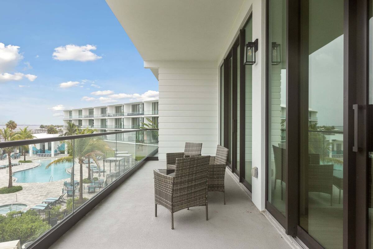 Een balkon met rieten stoelen, een glazen hek en uitzicht op een zwembad en hotelgebouwen.