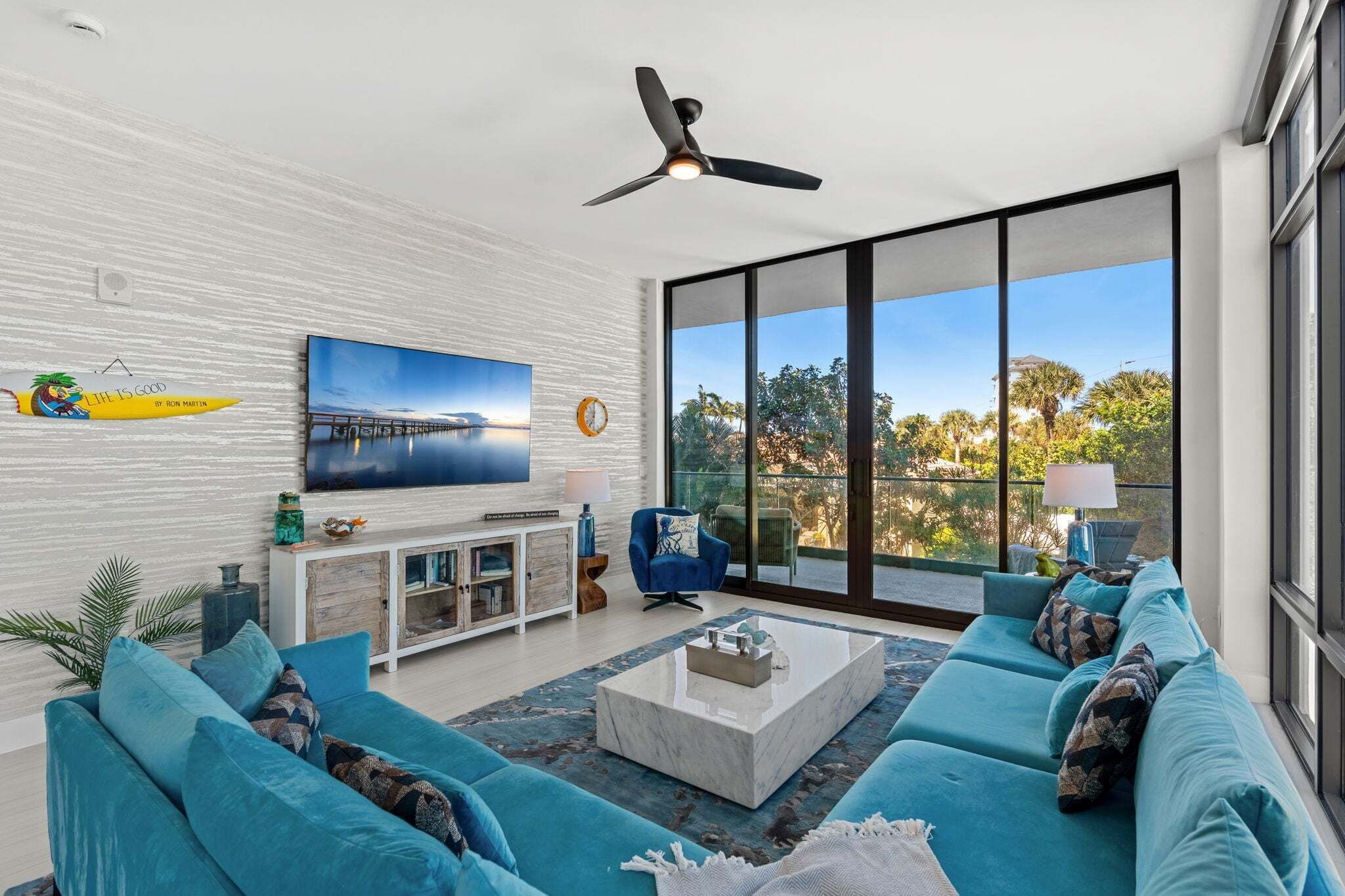 A modern living room with a large TV, a ceiling fan, and a view of trees through large windows.