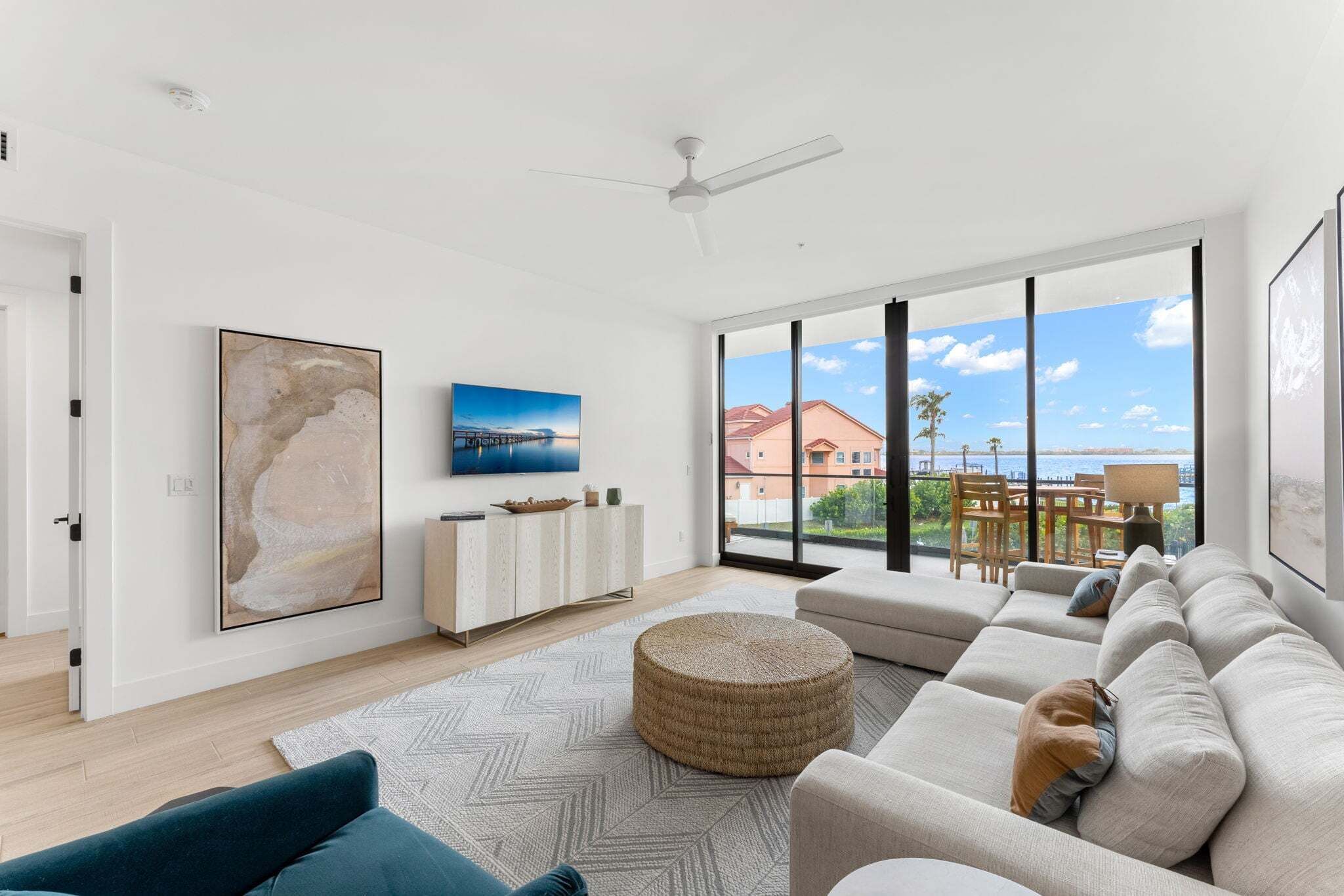 A modern living room with a large window, a ceiling fan, a sofa, a round ottoman, and artwork on the walls.