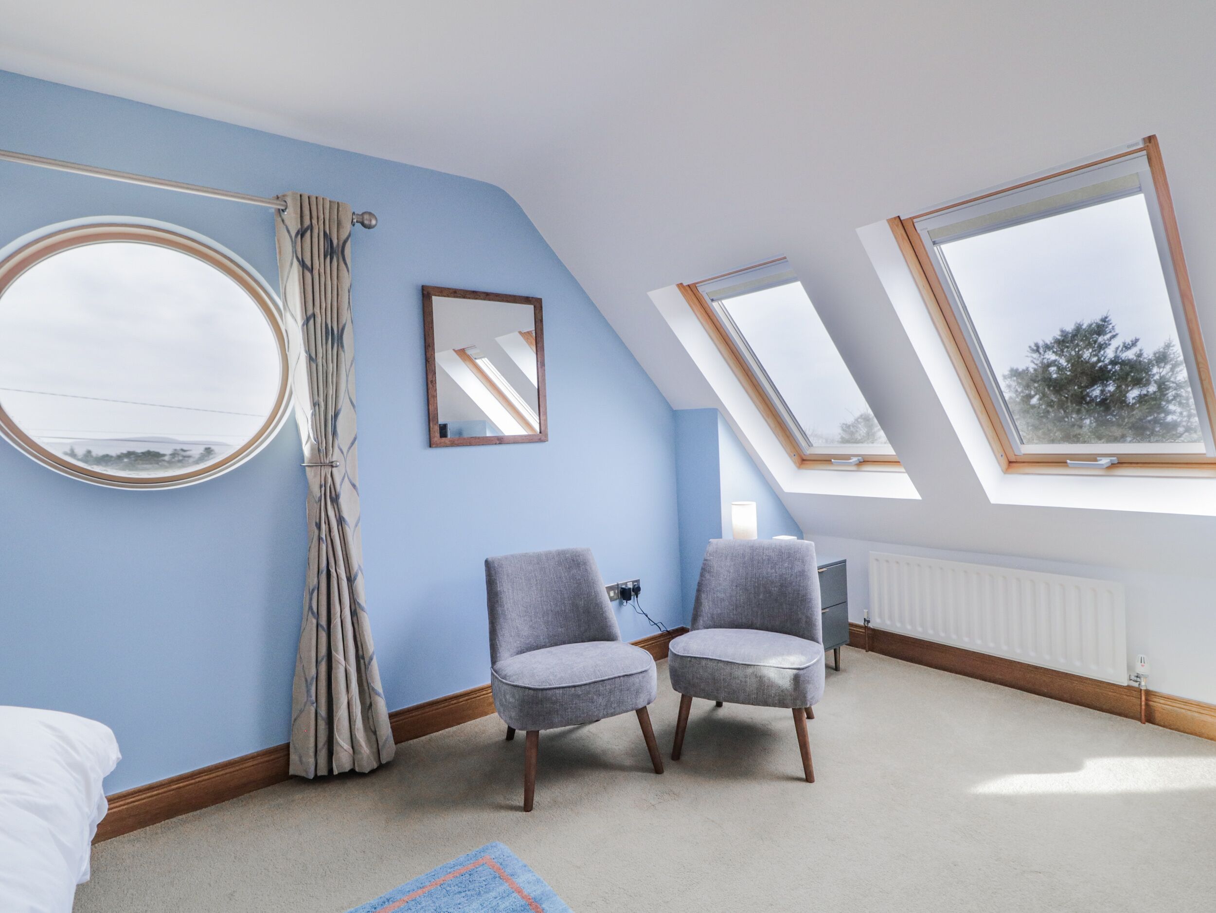 A bedroom with a skylight, two grey armchairs, a round mirror, and a window with a view of trees.