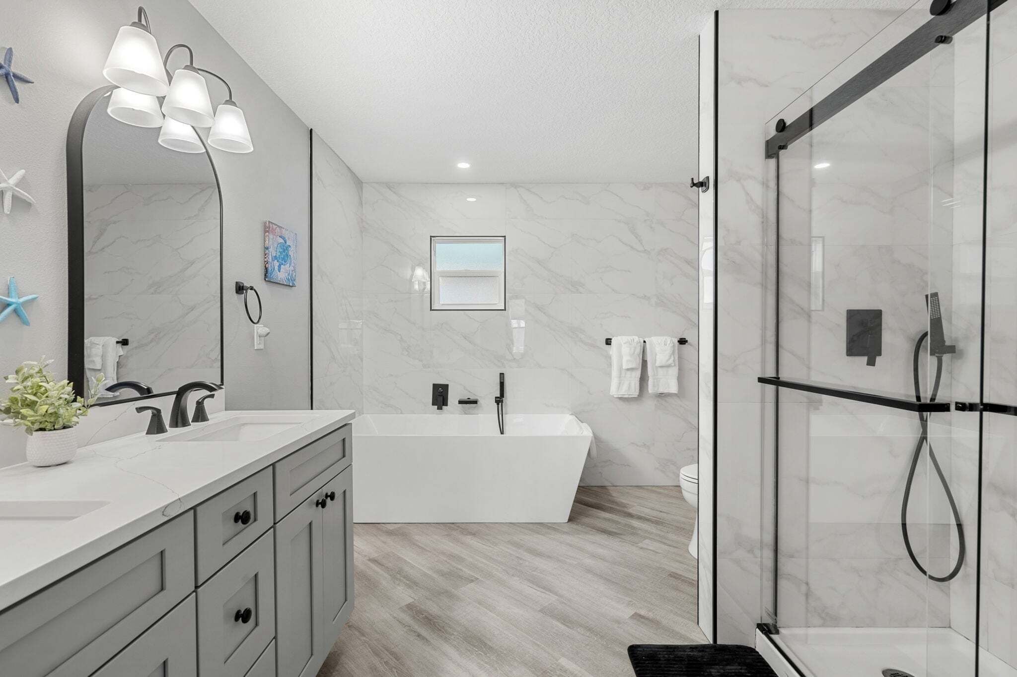 A modern bathroom with a large mirror, a freestanding bathtub, a glass-enclosed shower, and a wooden floor.