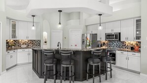 A modern kitchen with a central island, bar stools, white cabinets, and a tiled backsplash. - 218 Cherry Drive (Melbourne Beach)