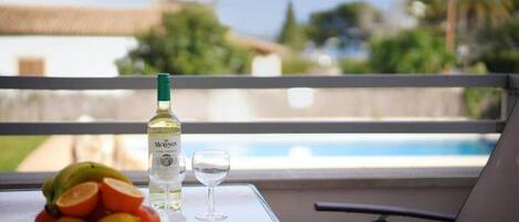 Una mesa con una botella de vino, una copa y un cuenco de fruta en un balcón con vista a una piscina y follaje.
