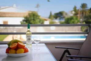 A table with a bottle of wine, a glass, and a bowl of fruit on a balcony overlooking a swimming pool and greenery. - Apartment Veleta (Port de Pollença)