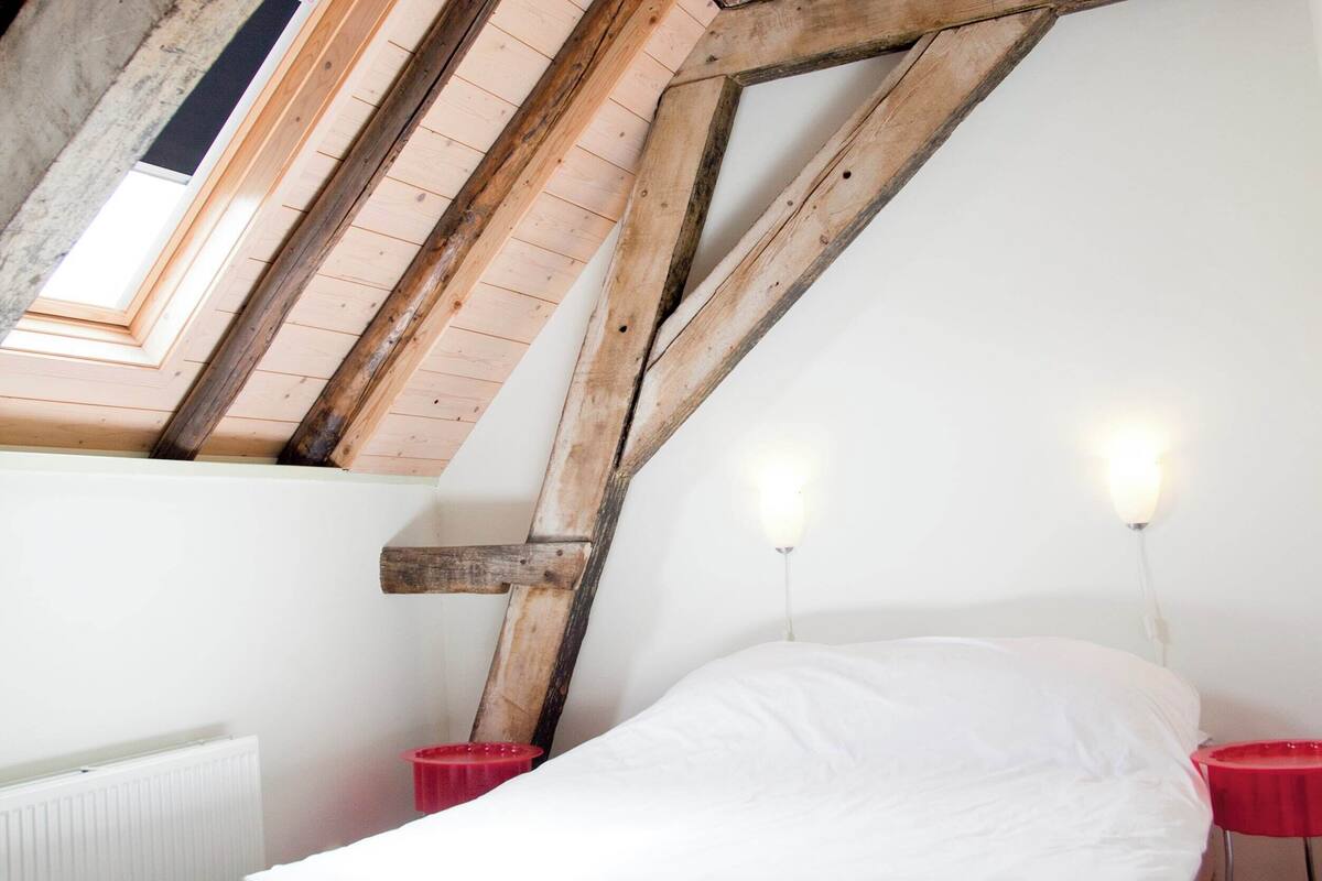 Een slaapkamer met een schuine kap, houten balken, een bed met witte beddengoed en twee nachtkastjes met rode lampen.