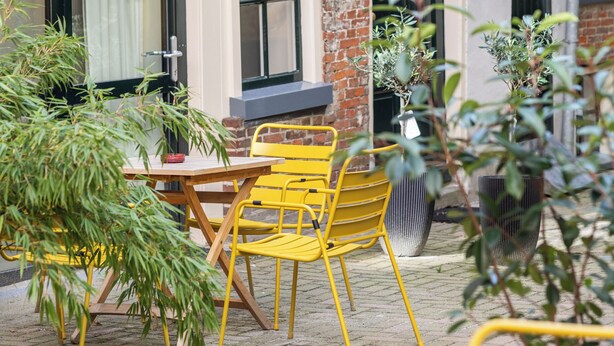 Een bakstenen gebouw met een raam waarop 'GELK INGE HOF' staat, en een binnentuin met gele stoelen en plantenpotten.