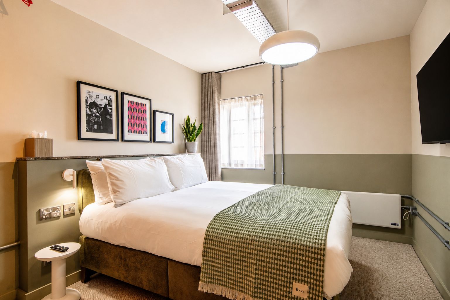 Habitación de hotel con cama, mesita de noche, lámpara, televisor montado en la pared y cuadros enmarcados.