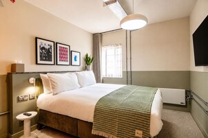 Habitación de hotel con cama, mesita de noche, lámpara, televisor montado en la pared y cuadros enmarcados.