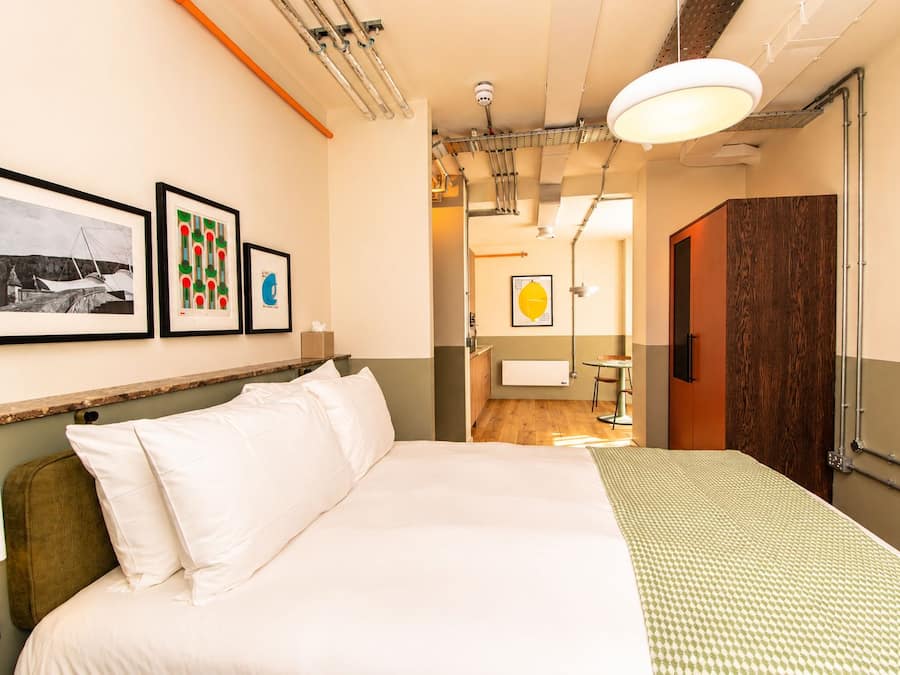 Une chambre d’hôtel avec un grand lit, une tête de lit en bois, des toilettes fixées au mur, une table de chevet avec une lampe et des œuvres d’art encadrées accrochées au mur.