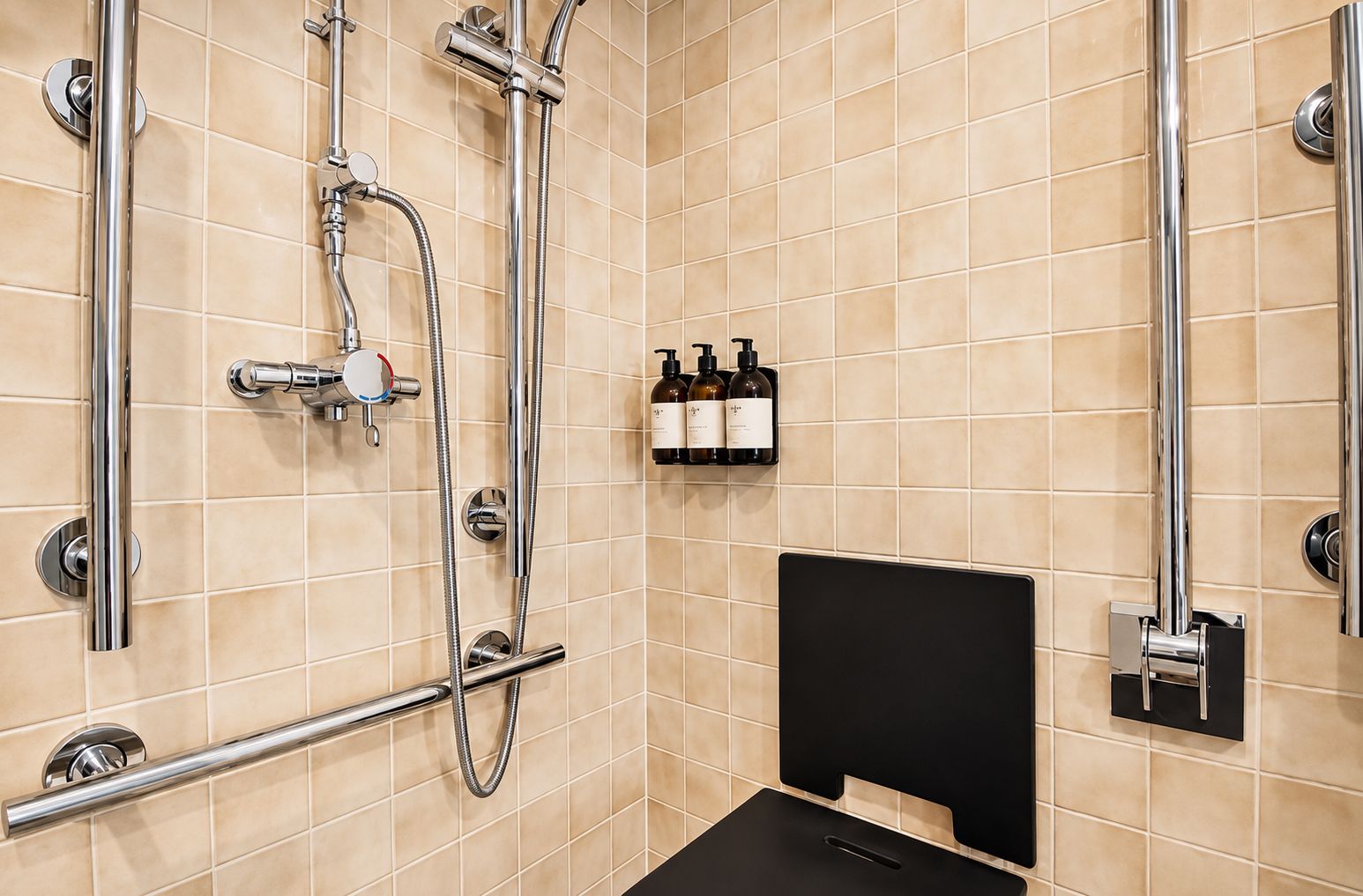 A modern bathroom with beige tiles, a black shower seat, and a set of toiletries on the wall.