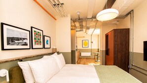 A hotel room with a large bed, a wooden headboard, a wall-mounted toilet, a bedside table with a lamp, and framed artwork on the wall. - Stay Hush (Edinburgh)