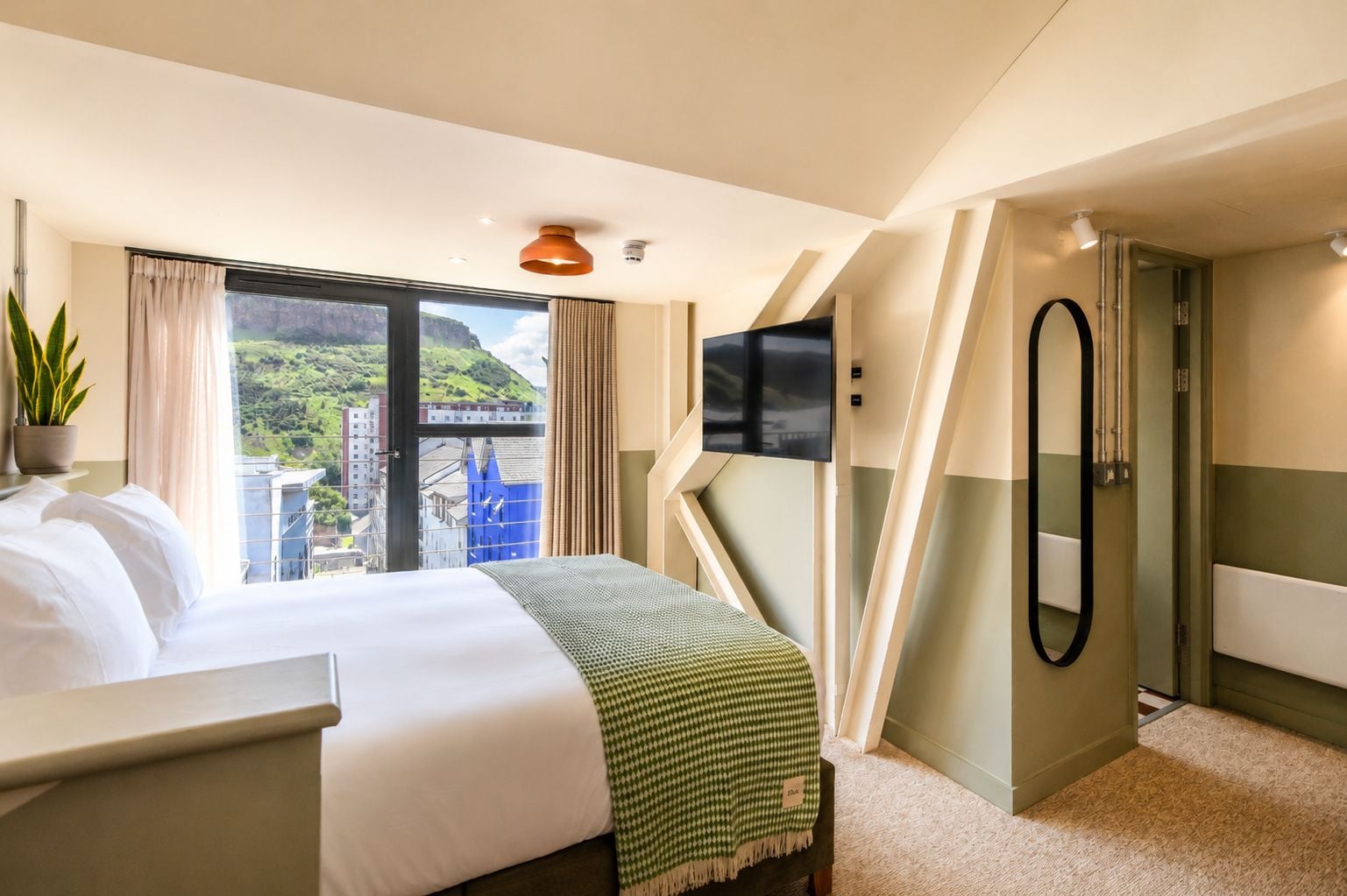 A hotel room with a bed, a TV, a mirror, and a view of a cityscape.