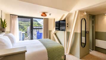A hotel room with a bed, a TV, a mirror, and a view of a cityscape.