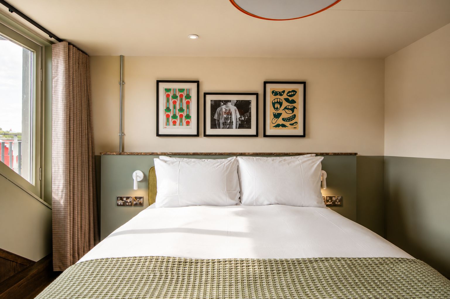 Hotelkamer met een groot bed, een hoofdeinde, een raam met gordijnen en drie ingelijste foto’s aan de muur.