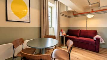 A room with a round table, two chairs, a red sofa, a small side table with books, and a framed artwork on the wall.