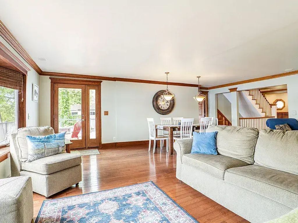 A spacious living room with a large sofa, a coffee table, and a dining area with a table and chairs. There is a staircase leading to an upper level.