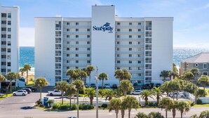 Een meerlaags hotel met een opvallend SeaSpray WEST bord, balkon en een parkeerplaats met auto's en palmbomen.