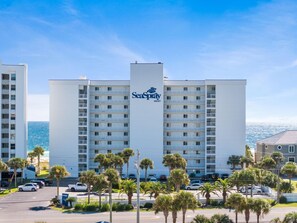 Ein mehrstöckiges Hotel mit einem gut sichtbaren SeaSpray WEST Schild, Balkonen und einem Parkplatz mit Autos und Palmen.