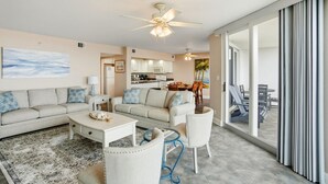 Een ruime woonkamer met een bank, salontafel en stoelen, een keuken op de achtergrond en een grote schuifpui die leidt naar een buitenruimte.