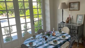 A dining area with a checkered tablecloth, white chairs, a lamp, and a view through a glass door to an outdoor garden. - A haven of peace just a few minutes from the sea, ideal for families (Montmartin-sur-Mer)