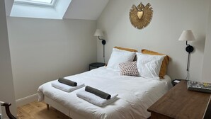A neatly made bed with white linens and two rolled towels, a wooden bedside table, a small round table, and a wall-mounted lamp. - A haven of peace just a few minutes from the sea, ideal for families (Montmartin-sur-Mer)