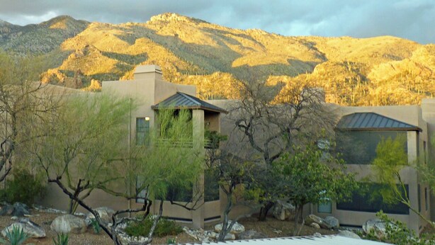 A modern building with a flat roof and large windows, surrounded by desert vegetation and mountains in the background. - Cozy 1-bedroom condo, in soothing Tucson with fitness room, pools & trails (Tucson)