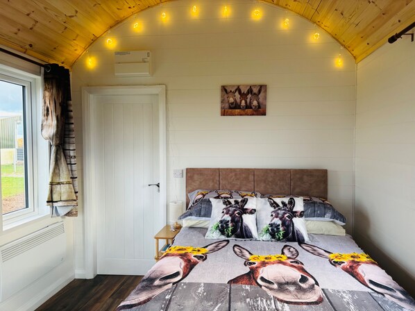 A bedroom with a wooden ceiling, a white door, a window with curtains, a bed with a floral and animal-themed bedspread, and a wall-mounted air conditioner. - The Dozy Donkey Shepherd Hut & Sauna Farm Stay (Ballyshannon)