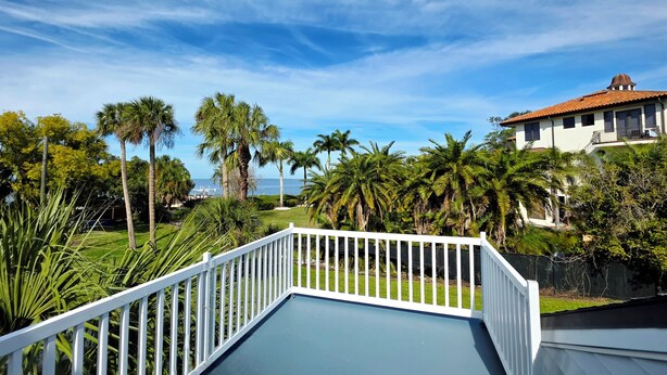Een balkon met een witte railing, met uitzicht op een verzorgd gazon en palmbomen, met een huis en de oceaan op de achtergrond.