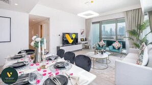 A modern living room with a dining area set for a meal. There is a flat-screen TV mounted on the wall, a sofa with decorative pillows, and a large window offering a city view. - Vogue | Downtown with Direct Access to Dubai Mall (Dubai)