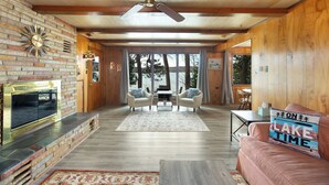 A cozy living room with a fireplace, wooden walls, and a view of trees and a lake. - Little Glen Lake 3 bedroom, 2 bath, Air Conditioning, Great Swimming & Fishing (Empire)