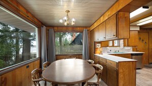 A wooden interior with a round dining table and chairs, a kitchen with wooden cabinets, and a large window with curtains. - Little Glen Lake 3 bedroom, 2 bath, Air Conditioning, Great Swimming & Fishing (Empire)
