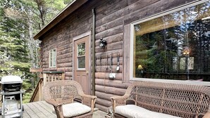 Eine Blockhütte mit Korbmöbeln auf einer Holzdiele, einem Grill und einem großen Fenster.