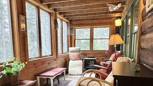Ein gemütliches Blockhaus-Interieur mit Holzwänden, großen Fenstern, einem Schaukelstuhl, einer Bank und einem Tisch.
