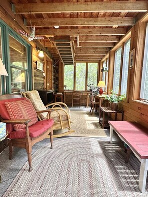 Ein gemütliches Blockhaus-Interieur mit Holzwänden, einem großen Fenster, einem Schaukelstuhl, einer Bank und einem Tisch mit Lampe.