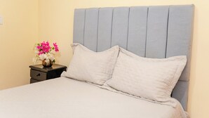 Ein Bett mit grauem Polsterkopfteil, ein Nachttisch mit Blumenvase und eine schlichte Wand.