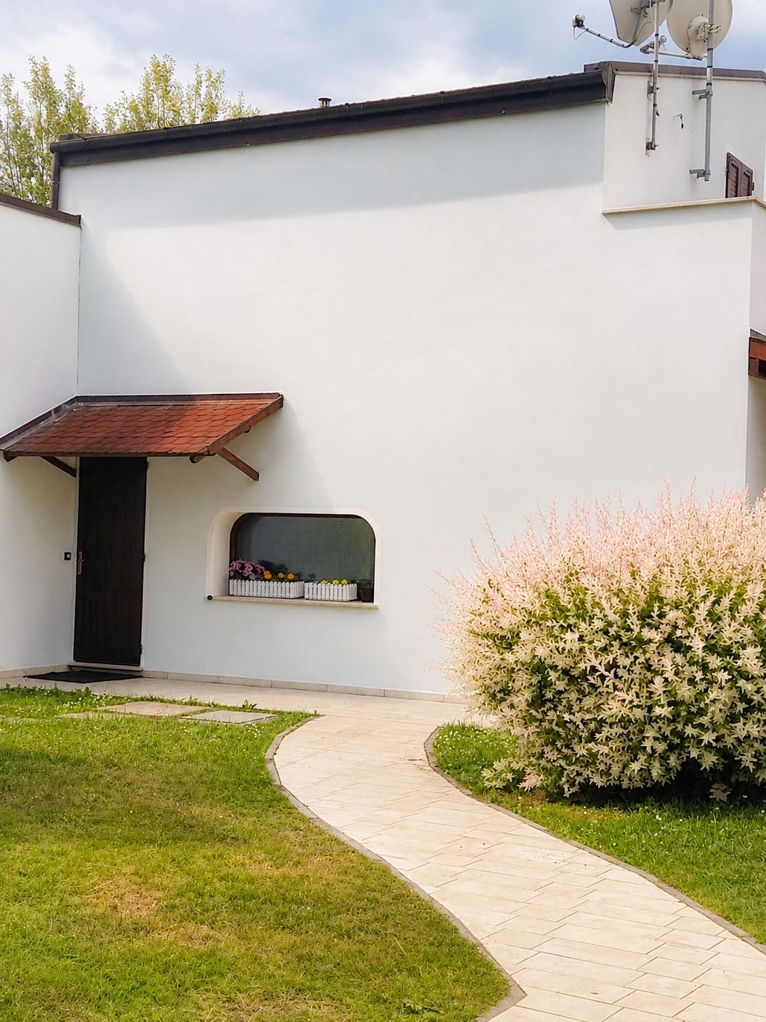Ein modernes Gebäude mit heller Fassade, einer dunklen Tür, einem kleinen Fenster mit weißem Rahmen und einem roten Ziegeldach über einer kleinen Veranda.