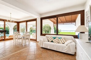 A spacious living area with a white sofa, a dining table with chairs, and a view of a swimming pool through large windows.